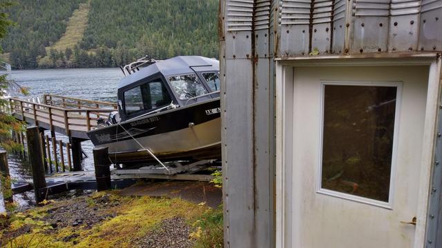A picture of the end of the boathouse (a metal shed, like a quonset) with a boat pulled up in front of it on the customized boat trolley (on a track); mid-background has dock and another boat; Alaskan sea inlet and opposite forested shoreline in distance