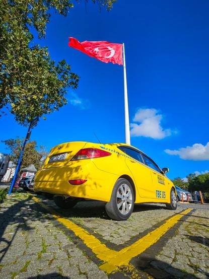 The image shows a bright yellow taxi parked on a cobblestone surface. Above the taxi, a large Turkish flag is waving on a tall flagpole against a clear blue sky. There are trees on the left side of the image, casting shadows on the ground. The taxi has a license plate visible, and there are other cars parked nearby.
