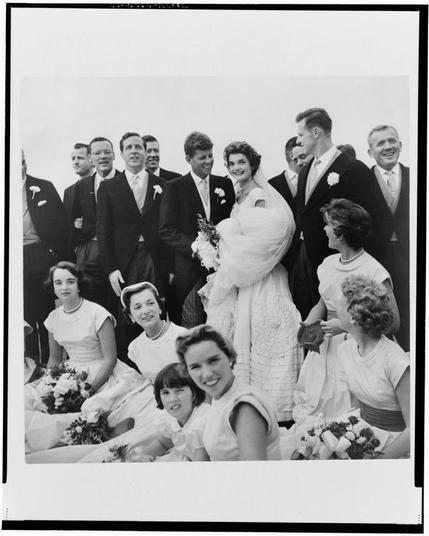 The image is a black and white photograph capturing a joyful moment of a wedding party. In the center stands the bride, Jackie Bouvier in an elegant dress with her arms crossed over her chest, holding a bouquet of flowers. Beside her are members of what appears to be both sides of the bridal party: groomsmen dressed in dark suits and boutonnieres on their lapels stand beside women wearing dresses; some seated while others pose standing or sitting around them. They all seem elated with wide smiles on their faces, showcasing a sense of camaraderie among themselves as they share this special occasion together.
