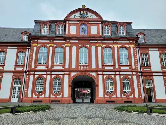 Eine barocke Fassade eines Klosteranbaus mit roten Kanten und Fenstereinfassungen und kleinen, weißen Flächen dazwischen. Im Giebel eine Marienfigur.