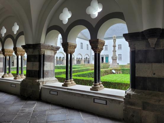 Blick aus dem romanischen Kreuzgang in den Innenhof mit einer barocken Marienstatue in der Mitte, die Hoffläche mit akkurat geschnittenen Hecken gefüllt. Die massiven Pfeiler des Kreuzgangs sind aus mehrfarbigem Gestein, dazwischen filigrane Säulen und reich verziete Kapitelle.