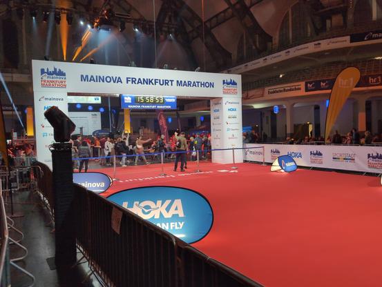 Das Ziel des Frankfurt Marathon in der Festhalle der Messe Frankfurt: ein weißer Torbogen mit großer Digitaluhr für die Bruttozeitnahme. Zum Torbogen führt ein roter Teppich, links und rechts abgezäunt. Im Hintergrund sind die Oberränge zu sehen, von denen aus der Zieleinlauf verfolgt werden kann.