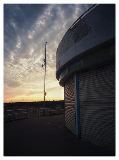 The setting sun forms a silhouette of a round fronted shop at the seaside. A lamp post stands tall in the distance