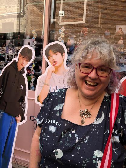 Person with fluffy hair, glasses, an octopus necklace, and a big delighted smile, standing in front of cardboard standees of Felix & Seungmin from Stray Kids. Seungmin is sucking on a lollipop.