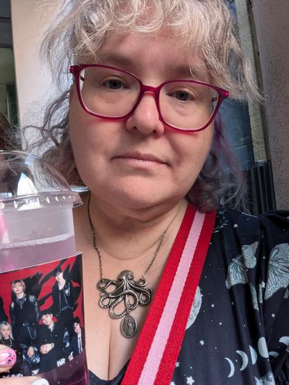 Selfie of person with fluffy hair and glasses holding up a cold drink with a stray kids cup sleeve
