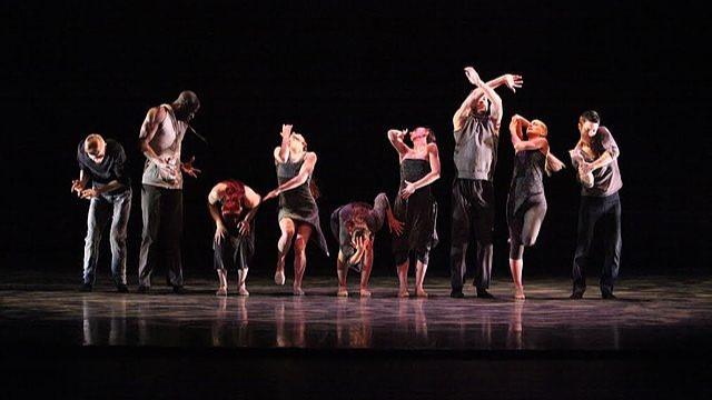 Nine dancers perform in a line on a dark stage