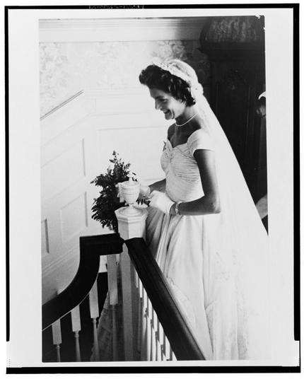In this black and white photograph, we see Jacqueline Bouvier in a wedding dress. She is standing on what appears to be the upper landing of an ornate staircase with balusters that have been polished over time by generations' hands before her own. The bride has dark hair pulled back into a bun from which a delicate veil cascades gracefully down and around one ear, adding elegance to her appearance.

Her attire consists of a sleeveless gown adorned with pleats on the bodice creating an intricate texture against the fabric's smoothness elsewhere. A subtle train trails behind her as she leans over slightly towards a small urn holding what appears to be flowers or greenery - perhaps part of her bouquet, which is not fully visible in this shot.

She holds a white vase containing more plants with both hands close together at chest level; one can almost hear the gentle clinking sound it makes against another. Her fingers are adorned with rings and she wears bracelets on each wrist enhancing her bridal look further. She gazes downwards towards these items, smiling softly revealing joyous anticipation for what lies ahead.

The background is minimally detailed but adds to the overall ambiance - a white wall behind her that reflects light creating an ethereal glow around her figure, while other decorative elements in view provide context of opulence and tradition fittingly complementing this moment captured in time.