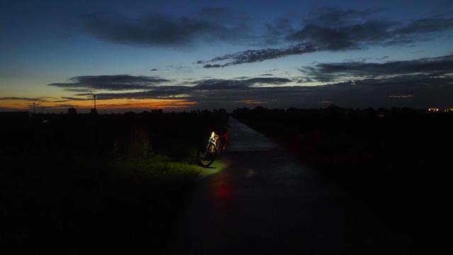 Beleuchtetes Fahrrad im dunklen auf einem Feldweg, im Hintergrund ist der Sonnenaufgang an einem Himmel mit ein paar Wolken,  zu erahnen