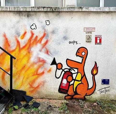 Streetart. A funny mural with a red dragon in comic style was sprayed/painted on a street wall next to a staircase. On the wall are signs for a heating oil valve and a fire extinguisher. The painted dragon stands next to it. It has a protruding lower jaw, a large and a small eye, circles for hands and a small flame burning on its tail. He is excitedly holding a fire extinguisher in his hands and trying to put out a sea of flames on the stairs. A big "Oops" is written above it and now we both know who started the fire.
Info: The anonymous artist "TocToc" is a French freelance illustrator and video artist. He exhibits his work in galleries around the world. In 2012, he created the simple figures DUDUSS when he was an art student and still sprays it on the walls of cities today.