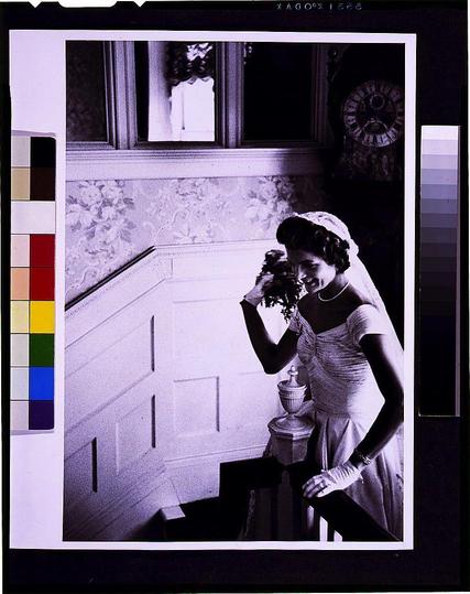 The image is a black and white photograph of a woman dressed in what appears to be bridal attire, with her gown featuring ruched fabric on the bodice. She stands near an ornate urn or vase that rests upon a ledge beside her. The setting seems elegant, possibly inside a historic building given the presence of patterned wallpaper behind her and a window reflecting light onto it.

The woman is smiling as she looks down at another object in her hand which isn't clearly visible but could be related to an event like a wedding or engagement due to her attire. A color chart with various hues can be seen on the left side, suggesting this image might have been part of a photo shoot involving different filters or lighting effects.

There is text above and below the photograph that mentions "KADOH," which may refer to a specific photographic process used during that time period as well as dates indicating when these types of photos were typically taken. The overall mood conveyed by this image seems joyous, capturing an intimate moment in what appears to be a significant life event for the woman depicted here.