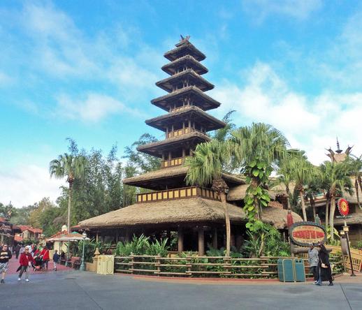 A multi-tiered, thatched roof structure resembling Asian architecture, surrounded by palm trees and lush greenery. The scene features visitors walking along a path, with a sign indicating the attraction "Enchanted Tiki Room" and two different designs of trash cans.