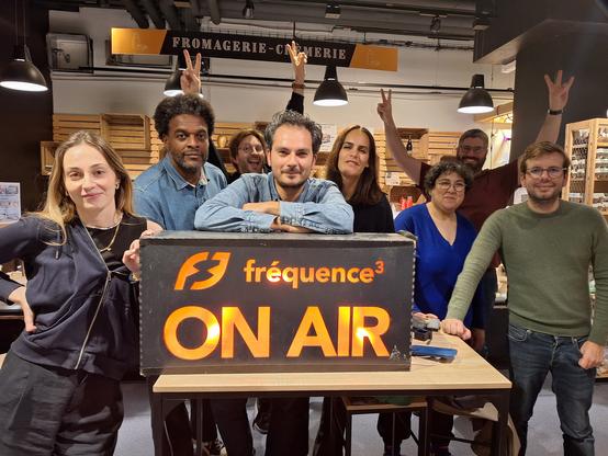 Photos de groupe, derrière un panneau lumineux orange "f3 fréquence3 ON AIR". 8 personnes sourient derrière, certains ont les bras en l'air et font des signes de la main. La photo est prise dans un magasin devant un rayon Fromagerie Crèmerie.