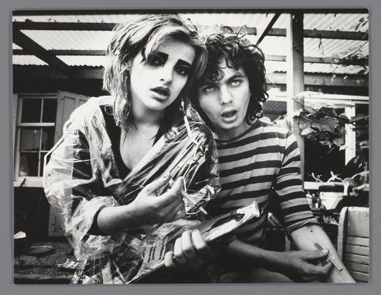 The image depicts two individuals in a candid, black and white photograph. The person on the left appears to be wearing makeup resembling that of a zombie or perhaps a reference to Frankenstein's monster, with dramatic eye makeup, darkened eyebrows, pale skin, and what seems like strands of hair styled to look messy or disheveled. They are also wrapped in a translucent plastic sheet, adding an element of decay or decayed state.

The person on the right is wearing a horizontally striped shirt and has curly, unkempt hair that appears tousled. This individual's expression could be interpreted as intense or serious, with wide eyes looking directly at the camera.

Both individuals are seated closely together; one holds what seems to be an open book or magazine in their lap while resting it on their thigh. The background is indistinct but suggests a casual setting with some plants and structures that may indicate they're outside near a residential area.

The photo has a raw, documentary feel typical of street photography from the late 20th century. It captures a moment between two people who might be friends or collaborators, possibly artists given their unique attire and makeup. The context suggests this image could have been taken during an informal photoshoot or while documenting cultural events in Amsterdam's Red Light District where Ed van der Elsken was based.

This photograph carries historical significance as it repres [...]