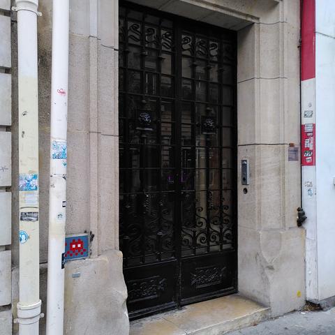 Une mosaïque bleue et rouge représentant un space invader, discrètement collé à côté de l'entrée d'un immeuble Haussmannien.