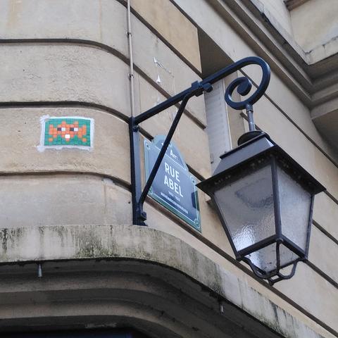 Une mosaïque verte et orange représentant un space invader, sur le mur d'un immeuble Haussmannien, à hauteur d'un réverbère.