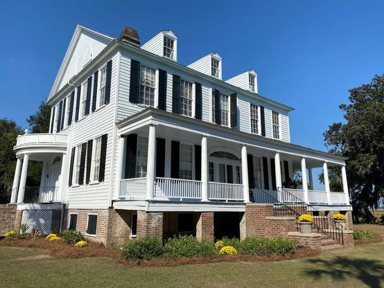Color image of a circa 1820 plantation home located in coastal South Carolina, USA. Blue sky above, image shows a two-story home on a brick foundation, dormer windows on the roof, side porches help up by pillars, with a staircase to the sea.