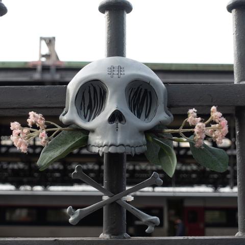 A front view photo of a stylized human skull,  cossbones sculpture and plastic flowers hanging on a train station fence . The skull is light gray with some black lines drawn on it, some artificial leaves and flowers are sticking out from each side of his upper jaw . it has no lower jaw. The skull has a train track drawn on the forehead.