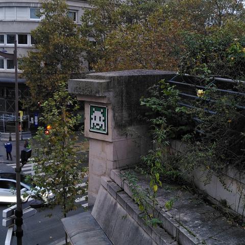 Une mosaïque blanche et verte représentant un space invader, collée sur un ancien pont ferroviaire, et entourée de verdure.