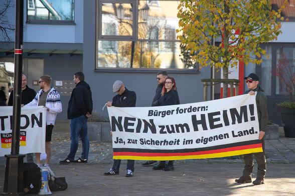 Mehrere Menschen stehen hinter einem Transparent mit der Aufschrift "Bürger fordern NEIN zum HEIM! - Ja, zur Sicherheit im eigenen Land" und einer Deutschland Fahne. 
Im Hintergrund ein Baum in Herbstfarben.
