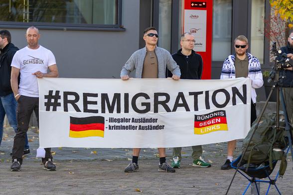 Mehrere Männer u.a. Daniel Kokott stehen hinter einem Transparent mit der Aufschrift:"#REMIGRATION - illigaler Ausländer - krimineller Ausländer - Bündnis gegen links" mit Deutschlandfahnen auf dem Transparent