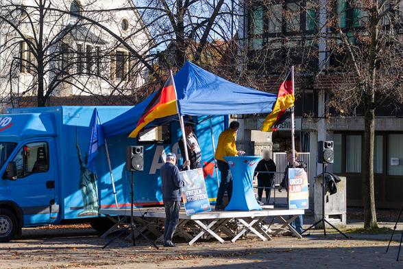 eine aufgebaute Bühne vor einem großen AfD Transporter in Blau mit Logos, der aufgebaute Pavillon fällt grade in Entstehen des Fotos nach hinten um. Auf der Bühne mehrere Männer