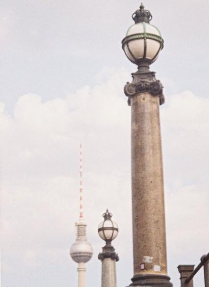 The Berlin Fernsehturm in the distance, two old street lamps imitate its shape in the front of the photo. The sky in the background is soft blue with creamy clouds.