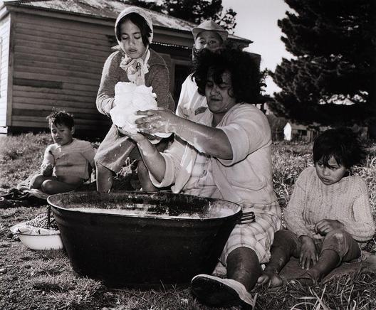 This black and white photograph captures a moment of domestic life involving several individuals, possibly family members. In the center, an adult appears to be engaged in washing or preparing fabric over a large bowl filled with water outdoors. This person is surrounded by others who are seated on the ground; some look towards the camera while others appear absorbed in their own activities.

The group consists of at least four adults and three children. One child sits near the individual working with the fabric, another rests close to them, possibly sleeping or resting, and a third observes from behind. The setting seems rural or semi-rural, suggested by the presence of trees, grassy ground, a wooden structure in the background that might be a shed or small cabin, and an overall lack of urban development.

The image evokes themes of simplicity, community living, manual labor, and perhaps hardship or resourcefulness given the outdoor setting for washing. It reflects on daily life practices possibly from past eras when such activities were more commonplace outside homes rather than within.