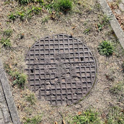 A round and outworn manhole cover with a pattern of small identical squares. The inscription is only partly readable and says "VEB KANALGUSS LUGAU" and something else.
