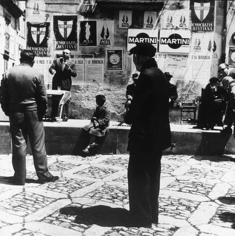The image is a black and white photograph depicting an outdoor urban scene with several people gathered on cobblestone pavement. In the foreground, there's a person standing and facing away from the camera, seemingly observing something out of frame to their right. This individual appears dressed in dark clothing.
Behind them, others are engaged in various activities: some sitting while talking or listening intently; one man stands addressing an audience with gesturing hands as if speaking passionately on a topic; another seated person is partially visible and seems more absorbed in the surroundings rather than participating in conversation.
The backdrop includes walls adorned with multiple posters featuring political messages, slogans such as "LIBERTAS," "DEMOCRAZIA CRIStIANA" (Christian Democracy), alongside names like MARTINI. The environment suggests a public square or market area where discussions and activities related to current events are taking place.