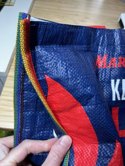 A close up view of the side of a Market Basket (local beloved grocery chain) insulated bag. The edge binding has been replaced with a strip of rainbow nylon webbing. A Caucasian hand is holding the edge so that both sides are visible.