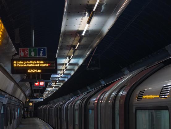 View along a London Undergound platform, with a tube stopped, doors closed. A departures display board has the time '02:24.18', and above it 'All trains go to King's Cross St. Pancras, Euston and Victoria'