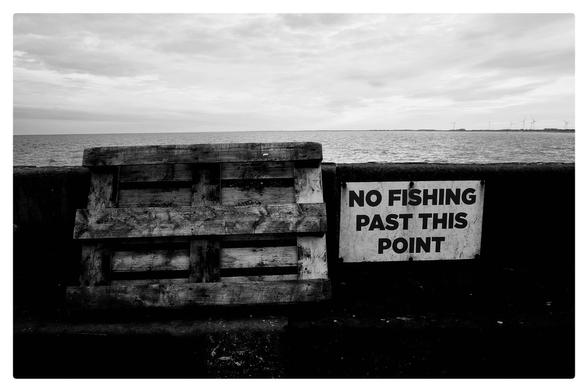 A no fishing sign on a sea wall next to a wooden pallet in high contrast black and white