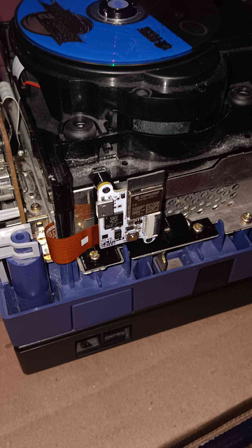 A Nintendo Gamecube with the top shell off, Sonic Adventure 2 in the Disk Drive, and a white modchip mounted on the inside connected to a brown ribbon cable coming out of the corner of the metal shielding the Disk Drive is mounted to