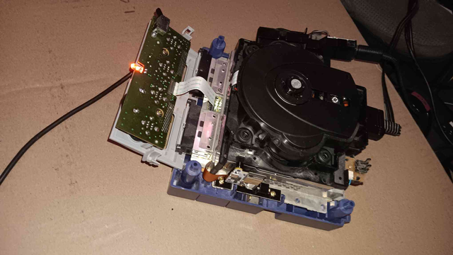 A Nintendo Gamecube with the top shell off. The disk drive is inserted, but the Fan/Power board and the Front IO boards hanging out and not attached. A controller, Power Cables, and AV cables are plugged in, and the orange power light on top of the front IO board is on