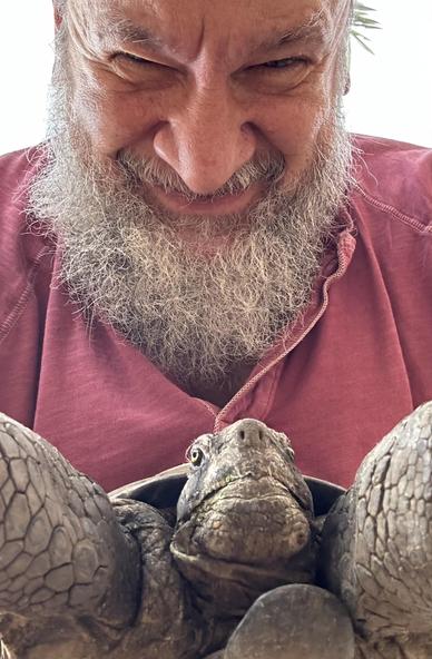 Smiling man with a gray beard and a red shirt holding a California desert tortoise with its front legs extended toward and behind the side edges of the photo