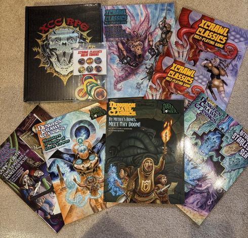 A pile of role playing game books, including the deluxe version of the Xcrawl Classics RPG and its game aids, plus Dungeon Crawl Classics RPG adventures such as Return to the Starless Sea and By Mitra’s Bones, Meet Thy Doom!