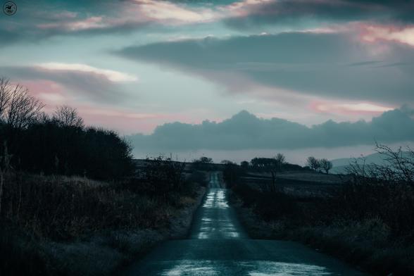 Long straight country road disappearing into the distance, with hedgerow and fields either side and cloudy sky above