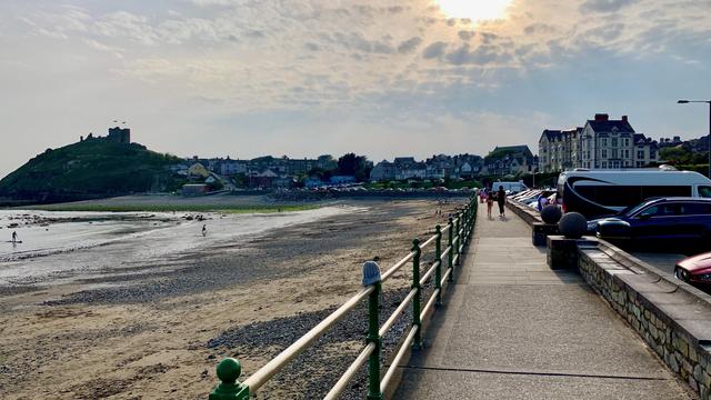 This photo captures a serene coastal scene. The foreground shows a paved walkway bordered by a green metal railing, with a stone wall along the side. Several cars are parked to the right of the walkway, while a few people are strolling along it, enjoying the view.

On the left side, a sandy beach with patches of pebbles stretches out toward the water, where a couple of people are visible, likely enjoying low tide. In the background, a hill rises with Criccieth Castle perched at the top. The structure flies two flags, giving it a prominent appearance against the sky.

The sun, partially obscured by thin clouds, creates a warm, diffused light, casting gentle reflections on the scene. The architecture of the buildings along the shore hints at a quaint, coastal town, likely in the UK or a similar region.