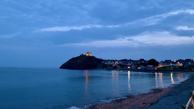 This image captures a peaceful coastal scene at dusk or early evening. The sky is a deep blue, with soft clouds adding texture. The sea is calm, reflecting the cool blue tones of the sky, while gentle waves lap against a pebble-strewn beach in the foreground.

A hill rises prominently on the left, topped by Criccieth Castle, which is illuminated, casting a warm glow against the surrounding blue. This warm light, along with the lights from buildings along the shoreline, reflects softly on the water, creating a serene and picturesque effect.

In the distance, the small town is nestled along the coast, with several lights visible in the houses, adding a cozy, inviting feel to the scene. This image beautifully captures the tranquility of a coastal town as day transitions to night.