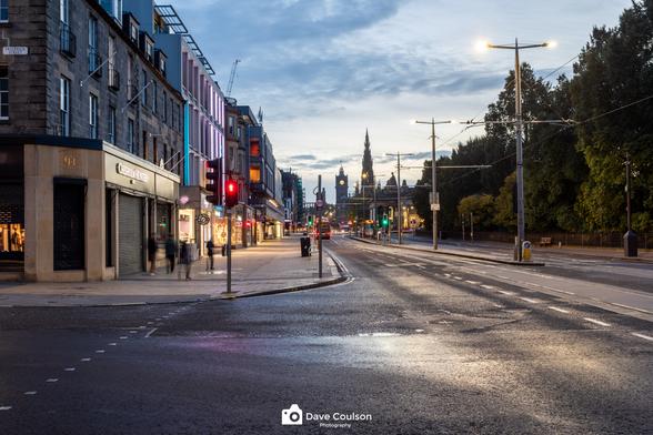 A view along Princes Street in Edinburgh early in the morning with lights from shop windows illuminating the street and Scott Monument and Balmoral Hotel in the distance.