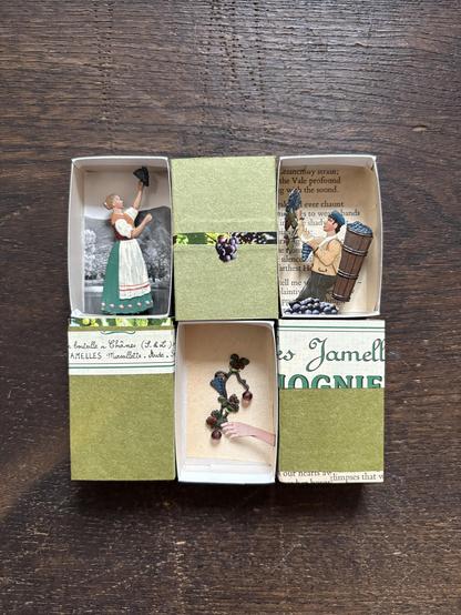 A wine label, moss green paper, and a page from a poetry book are used to cover the outer three matchboxes. One box features a tin ornament of a woman in traditional dress holding a grape, set against a black-and-white landscape photograph. Inside another matchbox, a small paper hand holds a decorative grape branch with purple beads. The third box displays a tin ornament of a man collecting grapes with a basket on his back, with a page from a poetry book as the background.