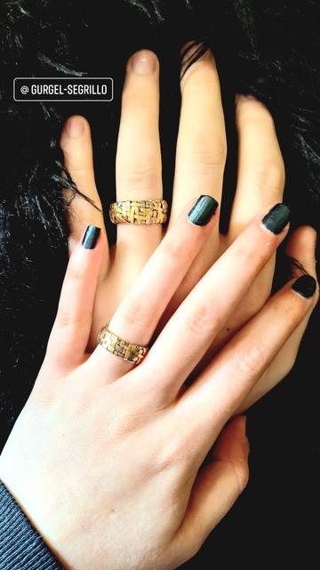 Two hands touching, one with black nail varnish, both wearing woven rings handcrafted with ribbons of yellow gold, over a black faux-fur background.