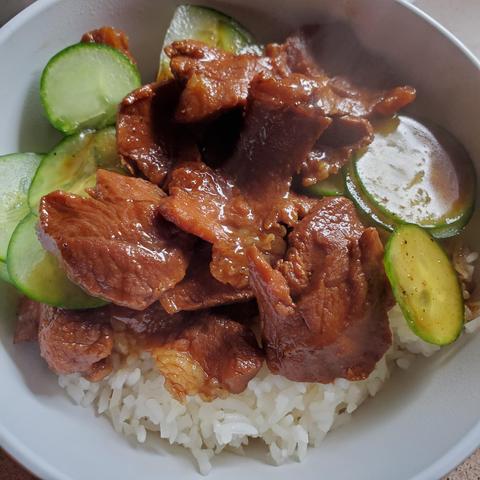 A bowl of steamed rice with sliced cucumbers & black pepper soy braised pork & braised sauce poured over.