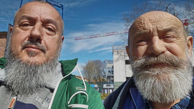 US...
The image shows two men with beards standing outdoors. They're in front of a clear blue sky with some trees and a building under construction in the background. The man on the left is wearing a green jacket, and the man on the right has a larger mustache and is wearing a dark coat.