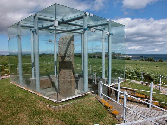The superb glass encased Shandwick Stone. The image shows a diagonal view of a large glass box wrapped around light blue-grey internal metal supports. This largely fills the left-hand two thirds of the frame. Inside it is a large Pictish carved stone with one highly decorated face visible. A fence surrounds the box and in the background the ground slopes away to the sea which is visible in the middle right of the frame. The scene is in sunlight.