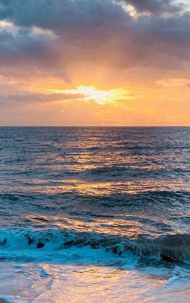 Color image of sunrise over the Atlantic Ocean, Edisto Beach, Edisto Island, South Carolina, USA. Image shows an active ocean at the waterfront, the clouds parting enoughto allow the sun to shine through over the sea as the waves crash the shore. Clouds above are white to blue to gray to orange as they surround the sun, with crepuscular rays shining all around, the sea's surface rough and tumble, with the sun's orange beams bouncing off the waves.