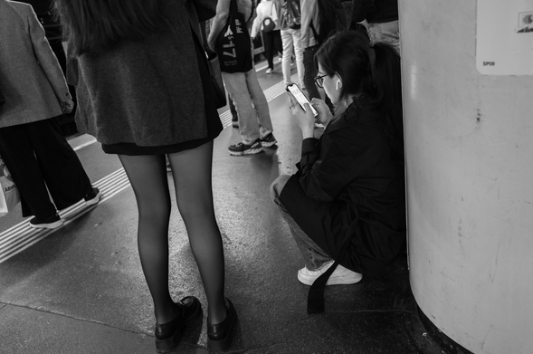 Black and white photograph taken in Vienna, showing a busy urban scene. A young woman is squatting by a column, focused on her phone with earbuds in, while another person stands nearby in tights and loafers. People walk around them in the background.