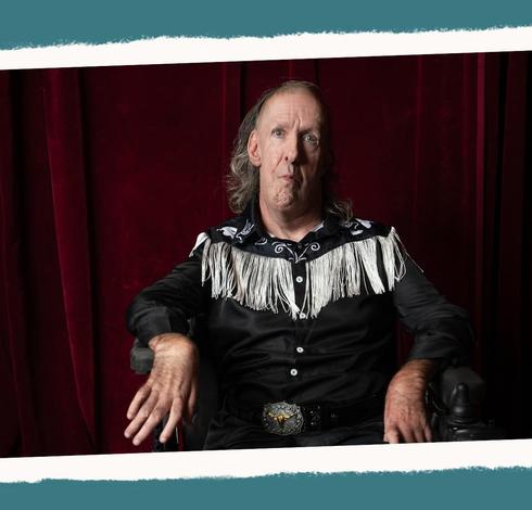 A man with long silver grey hair wearing a black western shirt with white fringe and a black leather belt with a big silver buckle, gazes at us as he sits in a wheelchair, a dark red curtain in the background.