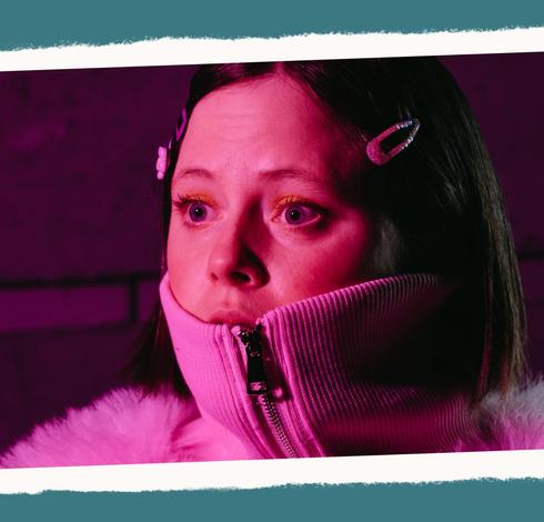 Close-up of a young person with a dark brown blunt hair cut, berettes at their temples, wearing a pink zippered top, the collar pulled up over their mouth, eyes wide in shock as they gaze to the left.
