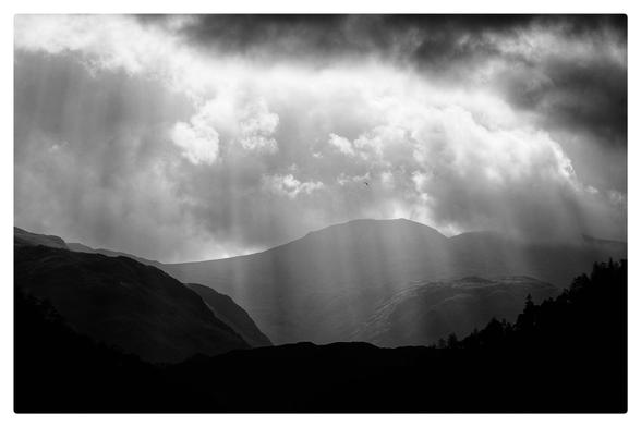 A silhouetted scene with sun rays bursting down from moody clouds over mountains.